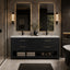 A modern double-sink bathroom vanity in a real bathroom setting, featuring a dark wood cabinet with gold handles, a white marble countertop, two rectangular backlit mirrors, wall sconces, folded towels, and a bathtub edge visible on the right.