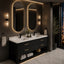Modern double-sink bathroom vanity with a black ribbed cabinet, white countertop, gold-tone handles, and two large oval mirrors backlit by wall sconces. The bathroom has dark textured walls, marble-look flooring, and a city-view window at night.