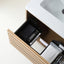 A bathroom drawer organizer set inside an open wooden vanity drawer, holding a black textured pouch, a white tube, and packaged items. The drawer is shown next to a white sink countertop.
