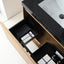 An open bathroom vanity drawer with a black organizer tray holding toiletries, shown next to a black countertop and white sink.