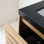 Close-up of a modern vanity cabinet with a glossy black countertop, light wood drawer fronts, and an open lower drawer showing storage space.