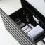 A modern dark gray drawer with horizontal slats, shown open to reveal a black interior organizer containing toiletries, a white towel, and a power outlet.