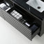 A modern black bathroom vanity drawer organizer shown open, containing white and gray bathroom accessories and toiletries, with a white sink visible on a black countertop.