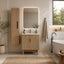 A modern bathroom vanity setup in a warm neutral room, featuring a light wood cabinet with woven cane doors, a white sink top, a matching tall storage cabinet, a backlit rectangular mirror, a hanging beige towel, and a freestanding bathtub nearby.