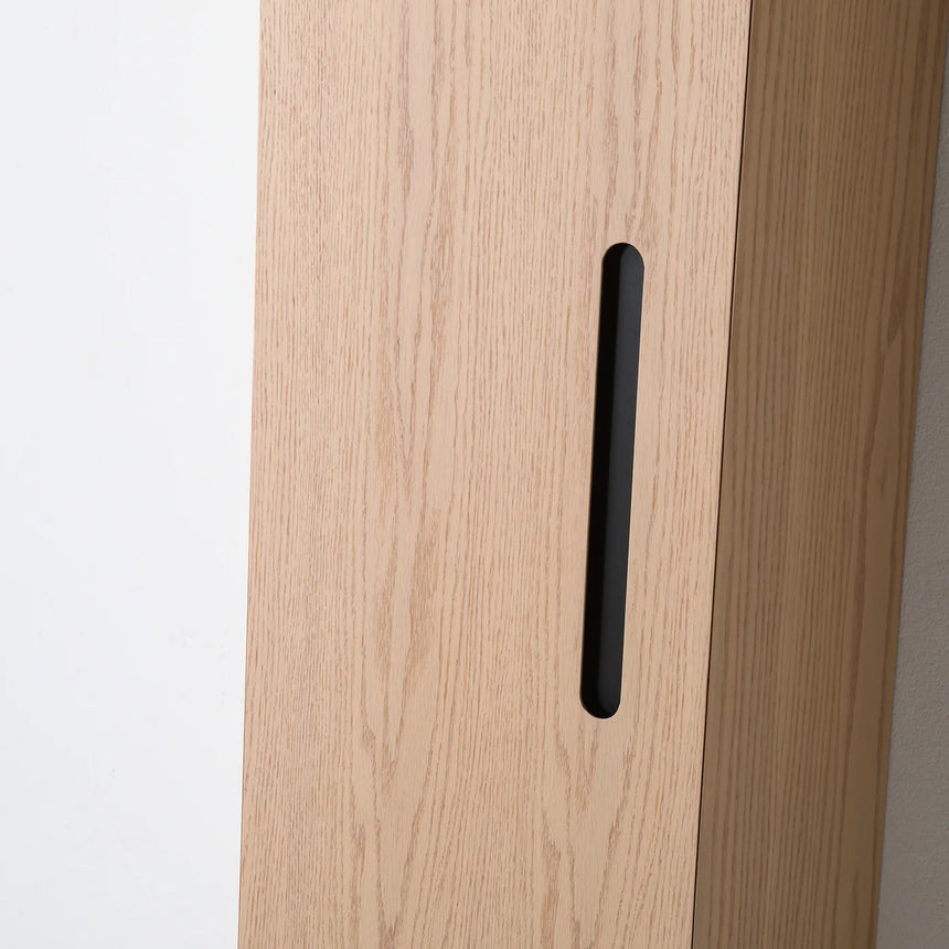 A tall, narrow wall-mounted cabinet in a light natural wood finish with a slim black vertical handle, shown against a plain light gray wall.