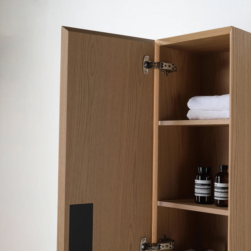 A tall, narrow wall-mounted cabinet in a light natural wood finish with a slim black vertical handle, shown against a plain light gray wall.