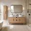 A modern bathroom vanity setup featuring a light oak wood double-sink cabinet with a white countertop, a large rounded rectangular mirror, a tall matching wood wall cabinet, and black hardware. The scene is styled with a glass shower enclosure, a textured rug, and warm wall sconces.