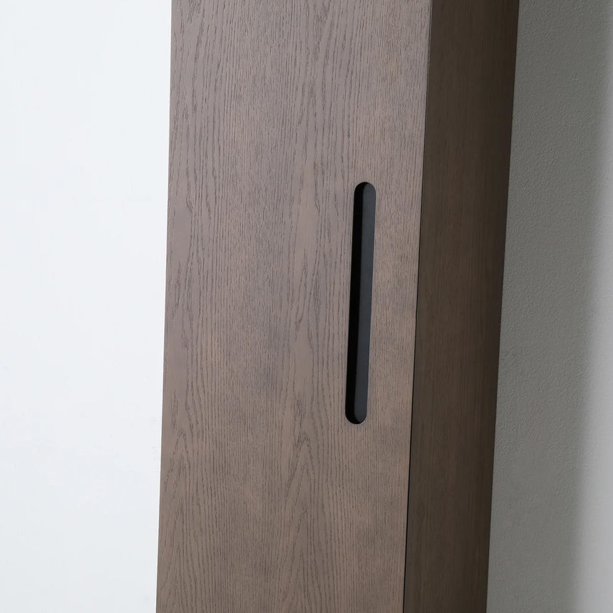 A tall, narrow wall-mounted cabinet in a light natural wood finish with a slim black vertical handle, shown against a plain light gray wall.