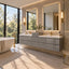 Modern double-sink bathroom vanity with glossy gray floating cabinets, white countertops, large backlit mirrors, and gold fixtures in a bright stone-tile bathroom with a freestanding tub and large windows.