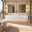 A modern bathroom interior featuring a glossy white double-sink vanity with two large mirrors, warm wall sconces, a freestanding bathtub, and a woven rug in a bright, sunlit room.
