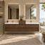 A modern floating double-sink bathroom vanity with a warm medium-brown wood finish and white integrated basins, set in a bright spa-like bathroom with large backlit mirrors, a window view, and a white freestanding tub nearby.