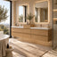 A modern double-sink bathroom vanity in a warm beige stone and light wood finish, featuring two illuminated rectangular mirrors, matte gold faucets, and a woven rug on a tiled floor.