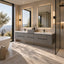 A modern bathroom interior with a double vanity featuring gray stone-look cabinets, white sinks, large rectangular mirrors with black frames, and warm wall lighting. A freestanding bathtub, glass shower enclosure, and large windows with a landscape view are visible.