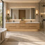 A modern double-sink bathroom vanity with light wood cabinets, white countertops, a large backlit mirror, and wall-mounted sconces, shown in a bright contemporary bathroom with a bathtub, rug, and glass shower enclosure.